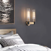 A contemporary bedroom showcases a wooden bed adorned with striped bedding and a textured gray pillow. Mounted on the dark gray wall is a sleek Morsale Palermo 2-Head Retro Brass Wall Sconce, providing warm lighting next to black and white abstract artwork.