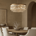A modern dining room features a round wooden table, cream upholstered chairs, and a decorative bowl with branches. The Bacci Crystal Chandelier from Morsale.com hangs above, casting warm light and enhancing the calm, elegant space with neutral tones.