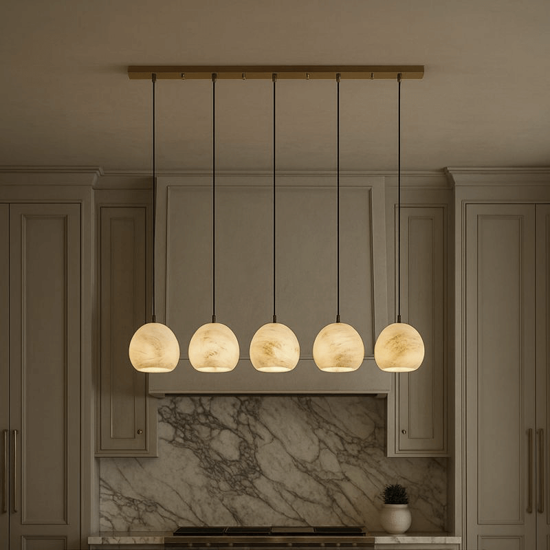 Auris 5-Light Linear Marble Chandelier by Morsale.com features adjustable suspension cords and dimmable LEDs. Its round, marble-patterned lights hang above a marble backsplash and countertop, complementing the modern kitchen’s light cabinetry.