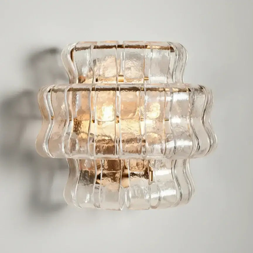 The Riviera Sconce by Morsale.com features a textured, wavy clear glass shade with warm light, creating an elegant, sculptural accent for any space—especially when mounted on a plain light-colored wall.