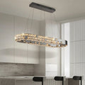 The Langley Dining Room Chandelier by Morsale.com with LED lights hangs above a kitchen island with black barstools, set against minimalist white cabinets, a gray backsplash, and a large window with vertical blinds.
