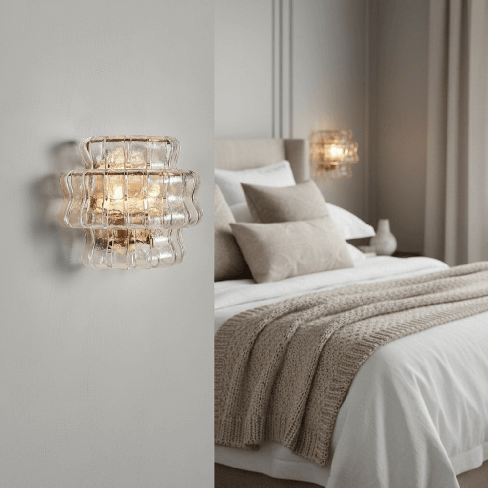 A modern bedroom with neutral tones showcases the Riviera Sconce by Morsale.com, casting warm light above a neatly made bed with beige and white bedding, textured pillows, and a knitted throw.