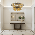 A bright foyer with white paneled walls, the Rivaro 2-Tier Brass Chandelier by Morsale.com, a gold-framed mirror, dark wood console table, and a large cream vase with green foliage atop polished marble floors.
