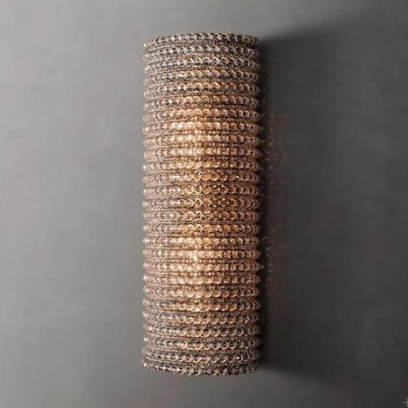 A cylindrical wall light fixture with a stainless steel frame is mounted on a grey wall. The Bergamo Crystal Wall Sconce by Morsale.com is adorned with numerous small, crystal-like beads, creating a textured, shimmering appearance. It emits warm, soft light from E14 LED bulbs that subtly highlight the intricate detailing of the beads.