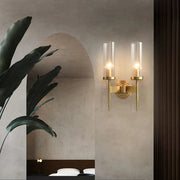 A modern interior features Morsale's Palermo Wall Sconce with dual cylindrical glass shades casting warm LED light. To the left, a large green plant adorns an archway to a bedroom with a neatly made bed and neutral decor, radiating modern elegance.