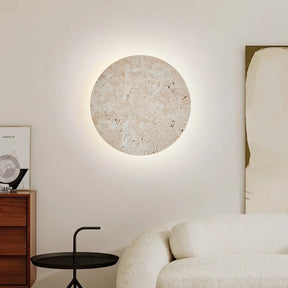 A modern, minimalistic living room features a Natural Travertine Wall Sconce from Morsale.com that emits a warm glow. Below the sconce sits a round black side table and wooden drawers with framed artwork. A white couch is positioned to the right.