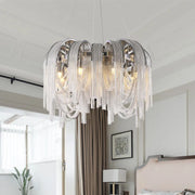 A modern bedroom features a large, sophisticated Cadena Chain Tassel Modern Chandelier from Morsale.com hanging from the ceiling. The chandelier has cascading strands of silver chain-like elements, encasing warm-toned E14 LED bulbs. The room includes a bed with a tufted headboard and large windows with sheer curtains.