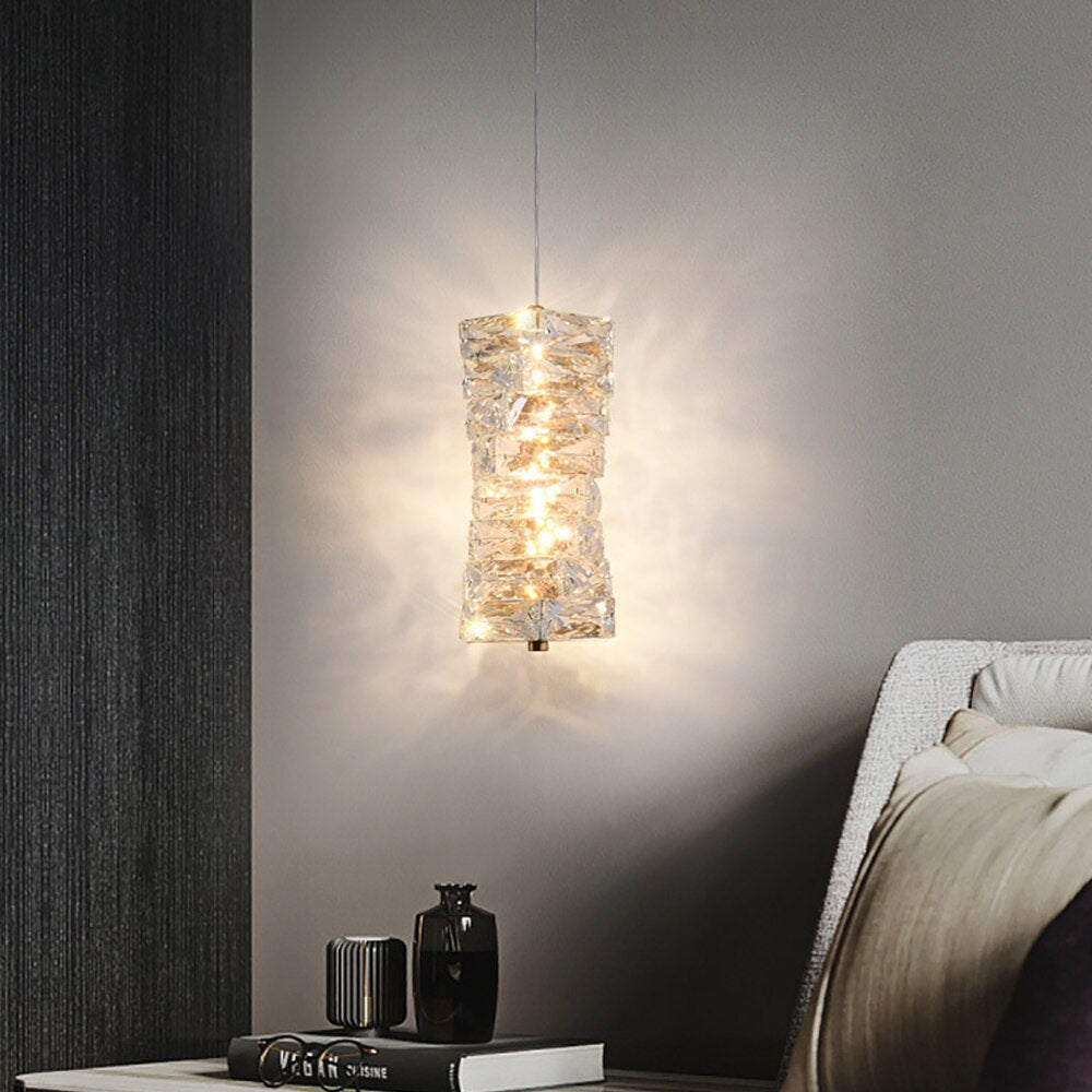 A modern Morsale.com Bacci Crystal Pendant Light Fixture emitting a warm glow hangs above a side table with books and decorative items in a dimly lit room with grey walls.