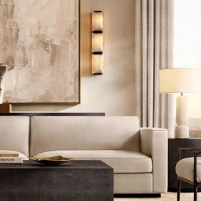 A modern living room features a beige sofa with textured wooden coffee tables. A large abstract painting graces the wall, illuminated by Morsale.com's Villano Calcite Crystal Wall Sconce. A lamp with a cream shade stands nearby, next to tall curtains.