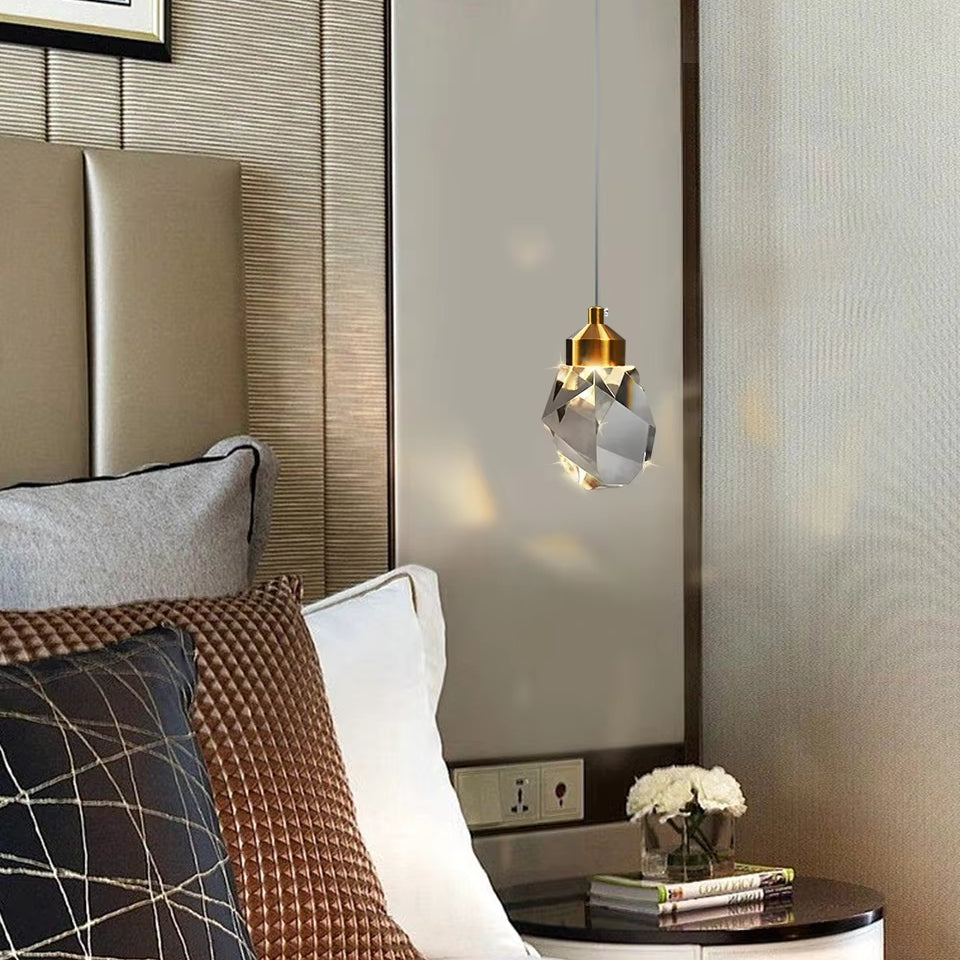 A modern bedroom features a cushioned headboard and decorative pillows in gray and brown hues. The Diamante Crystal Pendant Light by Morsale dangles gracefully beside the bed, enhancing the ambiance. On the small bedside table, a vase with white flowers pairs perfectly with a sleek digital clock, adding an elegant touch to the space.