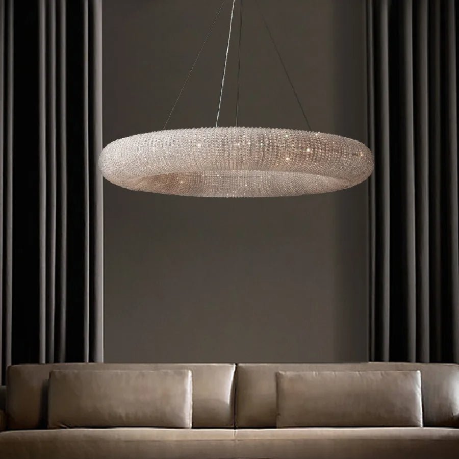 A modern living room features a large Bergamo Crystal Modern Chandelier from Morsale.com, with clear crystals hanging above a beige leather sofa. The light fixture boasts a textured, mesh-like surface and is suspended by thin cables, enhancing its luxurious appearance. The walls are dark, and two tall curtains frame the scene.