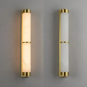 A Morsale 23" Natural Marble & Brass Wall Light featuring a vertical cylindrical design with a brass finish and energy-efficient LED technology. The light on the left is illuminated, casting a warm glow, while the identical fixture on the right remains off. Both are set against a gray background.