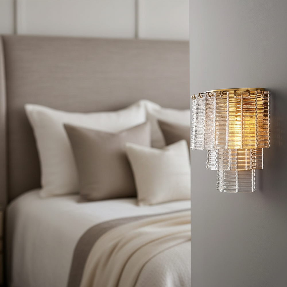 
                  
                    The Palermo Clear Crystal Wall Sconce by Chandelier Style, featuring gold accents, is lit beside a neatly made bed with neutral pillows and blankets in a modern, stylish bedroom.
                  
                
