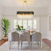 A modern dining room with a large, dimmable oval Verano Crystal Dining Room Light Fixture by Morsale.com featuring crystal accents and a gold frame. Six grey upholstered chairs surround a rectangular white table adorned with a green plant centerpiece and two white vases. A potted plant is in the corner near a sunny window, adding to the contemporary design.
