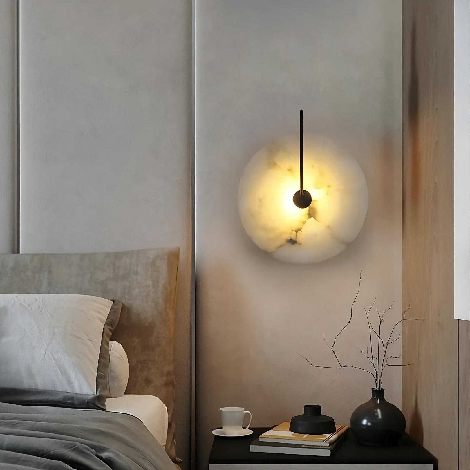 A modern bedroom features a soft gray bed with white and gray bedding. The room is illuminated by the Moonshade Natural Marble Sconce Light from Morsale.com, showcasing its unique texture above a minimalist black nightstand. The nightstand holds a small vase with twigs, a closed book, and a white coffee cup.