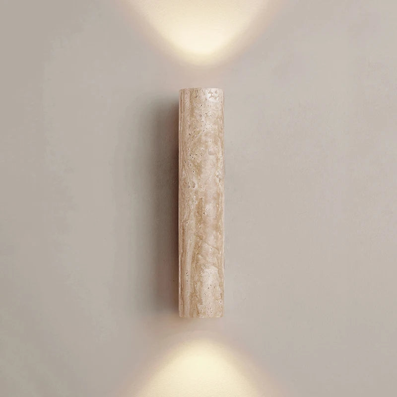 A vertically mounted **Natural Travertine Wall Lamp** by **YEBMLP** with a textured, light stone finish and champagne glass accents casts a soft, warm light both upwards and downwards, creating an ambient illumination effect on the light gray wall behind it.