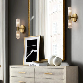 A stylish interior features a modern dresser with gold handles, topped with two white vases and a framed black-and-white abstract art piece. Two Morsale Palermo 12" Brass Wall Light Sconces with elongated glass shades flank a tall mirror reflecting part of a window and building facade, adding to the sophisticated elegance.