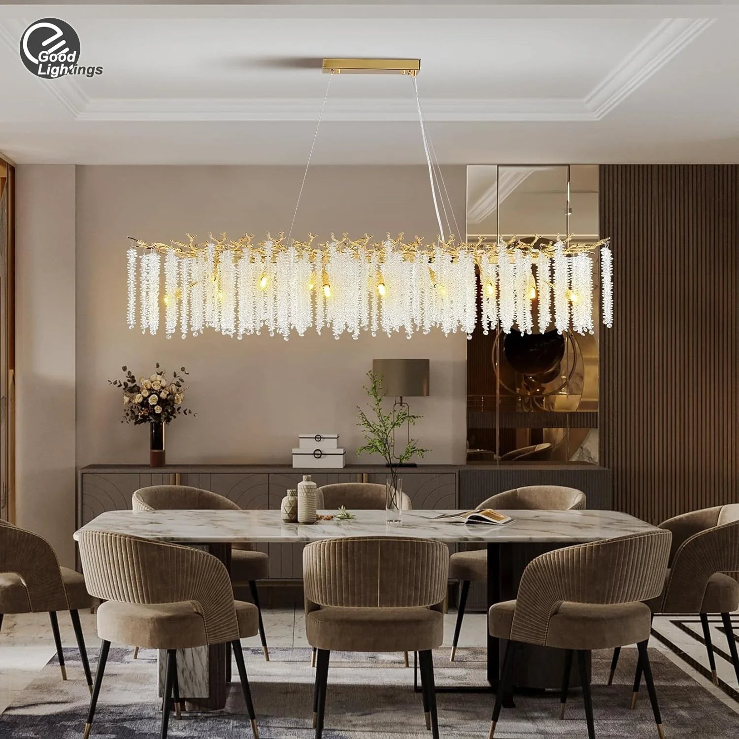 A modern, rectangular Albero Kitchen Ceiling Light Fixture from Morsale.com hangs above a dining table, featuring cascading crystal-like elements and multiple light bulbs. The room has a minimalist design with neutral tones, and a bowl of fruit can be seen on the table below the chandelier.