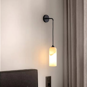 The Moonshade Natural Marble Wall Lights by Morsale.com feature a minimalist design with a black metal fixture and cylindrical frosted glass shade, emitting soft, warm light. Ideal for enhancing home decor, they perfectly complement dark-colored headboards and beige curtains on light-colored walls as an elegant wall sconce option.