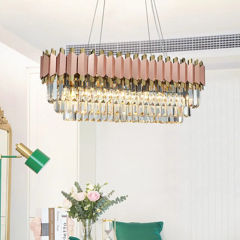 A modern rectangular dining room chandelier with gold and pink accents, the Gio 39" Rose Gold Dining Room Chandelier by Morsale.com, hangs from the ceiling. It features multiple crystal prisms and dimmable LED lights, illuminating the area. Below it, there's a green desk lamp, a vase with pink roses and eucalyptus, and a cozy green pillow on a chair or sofa.