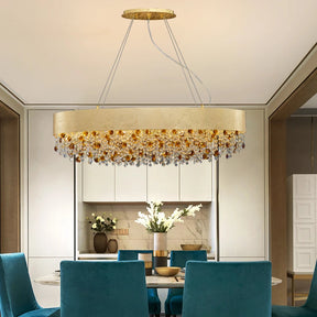 The Avani Crystal Dining Room Chandelier by Morsale.com is a modern luxury chandelier with a sleek, circular design. It features an arrangement of amber and clear crystals and gold accents hanging below a wide, gold band. The chandelier is suspended from the ceiling by thin wires, set against a plain gray background.