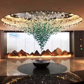 A modern lobby showcases luxury lighting with a Piovere Multi Pendant Chandelier from Morsale.com, resembling a waterfall of green and white crystals. Below, a circular dark fountain sits on marble flooring against frosted glass featuring a mountain landscape design.