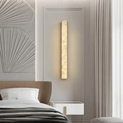A modern bedroom features the Moonshade Collection Marble Sconce in Black by Morsale.com, emitting warm light in a sleek vertical rectangle design. To the left, an upholstered headboard partially appears against textured wall art with a fan-like design. A small white bedside table is positioned next to light-colored curtains.