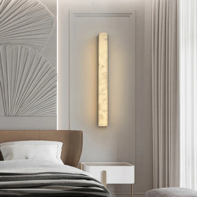 A modern bedroom features the Moonshade Collection Marble Sconce in Black by Morsale.com, emitting warm light in a sleek vertical rectangle design. To the left, an upholstered headboard partially appears against textured wall art with a fan-like design. A small white bedside table is positioned next to light-colored curtains.