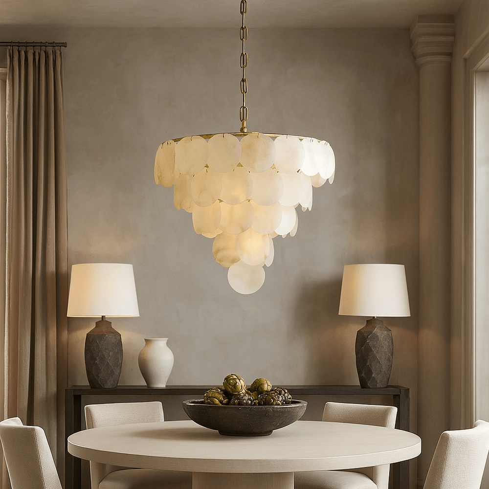 A round dining table with four chairs sits beneath the Porto Marble Modern Chandelier by Morsale.com. Behind, a sideboard displays two matching lamps, a vase, and a bowl against a neutral-toned wall with drapes.
