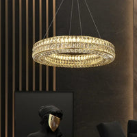 The Morsale Hutton Crystal Chandelier glows warmly above a dark-toned room, where a black geometric sculpture with a gold face mask and a matching black chair evoke an atmosphere of luxurious decor.