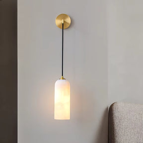 The Moonshade Natural Marble Wall Lights from Morsale.com feature a minimalist design with a gold circular base and a long black cord that suspends a cylindrical frosted white shade. These lights resemble elegant wall sconces, casting a soft glow beside the neutral-toned upholstered headboard, enhancing your sophisticated home decor.