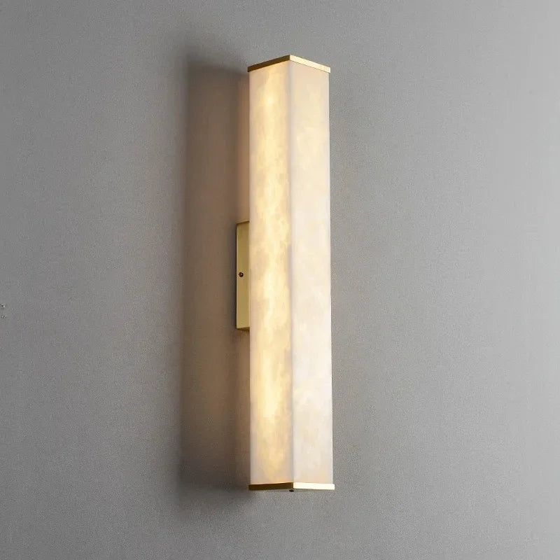 A modern wall-mounted light fixture with a sleek rectangular design. The Natural Marble Indoor Wall Sconce Light by Morsale.com, encased in frosted glass or acrylic cover, emits a soft, warm glow. It is mounted vertically on a plain gray wall.