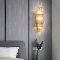 The Morsale.com Gio Crystal Wall Sconce, with a gold geometric frame and clear crystals, emits warm dimmable light on a gray paneled wall beside a bed with a gray headboard, white pillow, and a small plant on the nightstand.