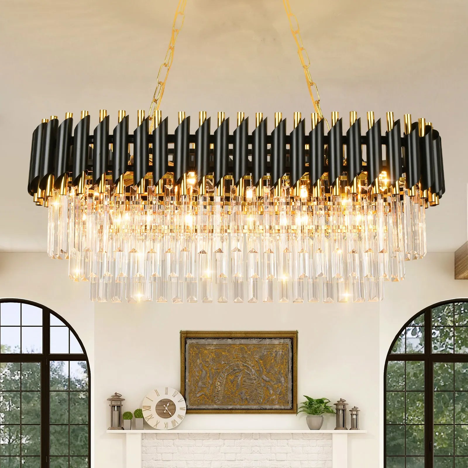 An elegant Verano Crystal Dining Room Light Fixture from Morsale.com with multiple tiers of cylindrical glass pieces hangs from a gold chain in the center of a room, exemplifying contemporary design. The dimmable chandelier features a mix of black and gold elements. Below, a white mantelpiece with decor items sits against a wall with two tall, arched windows.