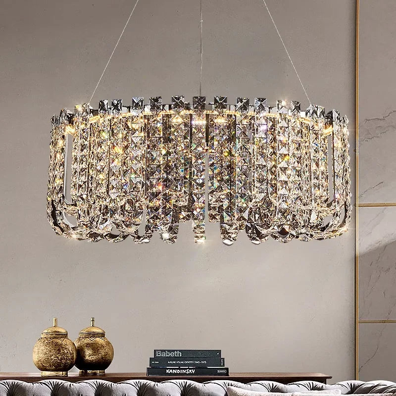 The Bacci Crystal Beads Chandelier by Morsale, featuring an intricate design and sparkling crystal accents, hangs gracefully from the ceiling, casting a luxurious glow. Below, a table adorned with decorative vases and a stack of books rests against a neutral-colored wall.