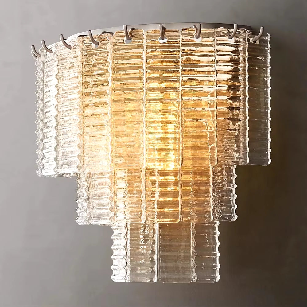 The Palermo Clear Crystal Wall Sconce by Chandelier Style is a wall-mounted light fixture with layered transparent glass panels that emit a warm, glowing light against a gray background. Its ridged texture adds luxurious crystal accents, resulting in a sophisticated and modern design.