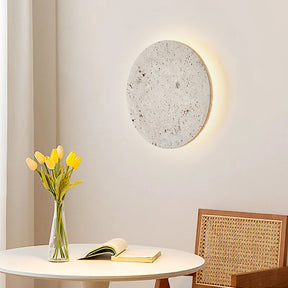 A modern minimalistic room features a round, Morsale.com Natural Travertine Wall Sconce emitting a soft glow. Below, a small table holds an open book and a vase with yellow tulips. Next to it stands a chair with a wicker back, all set against a light-colored wall and curtains.