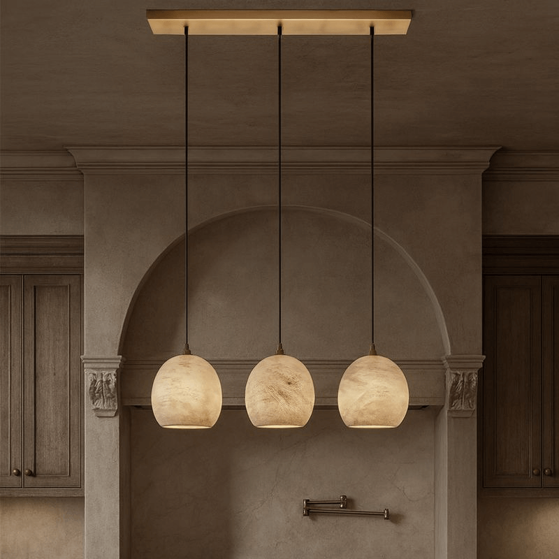 The Auris 3-Light Linear Marble Chandelier by Morsale.com features three globe pendants on adjustable cords, suspended in a row from a rectangular mount, adding ambient light to a neutral kitchen with wooden cabinets.