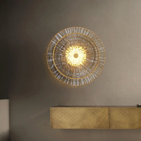 A circular Modern Glass Wall Sconce by Morsale.com with a radiant, sunburst design emits warm light. The handmade sconce features an intricate pattern and gold accents. Below it is a sleek, wall-mounted console with a textured, natural marble finish blending seamlessly with the neutral-toned wall.