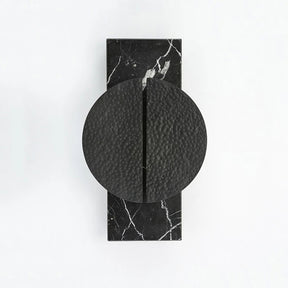 A modern, black Medieval Marble Wall Light Sconce by Bigman with a rectangular base and a circular handle made from textured metal, set against a white background. The sconce features a vertical groove down the center. The black marble base, reminiscent of an art deco wall sconce, boasts elegant white veining.