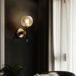 A Morsale Palermo Wall Sconce Light with a ribbed, textured glass globe shade is mounted on a dark wall next to floor-length curtains. The lamp's brass finish emits warm light, illuminating the surrounding area. A part of a fur-covered chair and a plant are visible at the bottom.