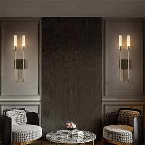 In a stylish sitting area, two patterned armchairs and a round marble coffee table adorned with books and a vase of flowers are beautifully set against a textured dark wall. The interior design lighting is elegantly enhanced by the Candela Contemporary Wall Sconces from Morsale, featuring sleek cylindrical lights.