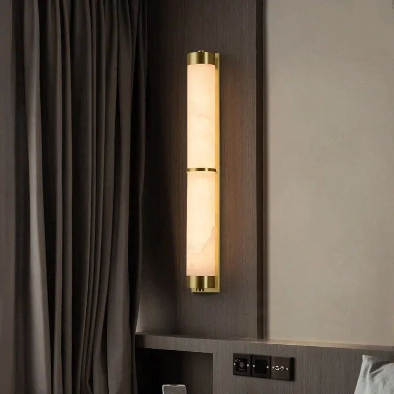 A modern, energy-efficient 23" Natural Marble & Brass Wall Light by Morsale with a vertical design is mounted on a gray wall. The sconce features two cylindrical lights separated by a brass accent, emitting a warm glow. Nearby, a dark curtain hangs from the ceiling and a section of a light-colored bed is visible.