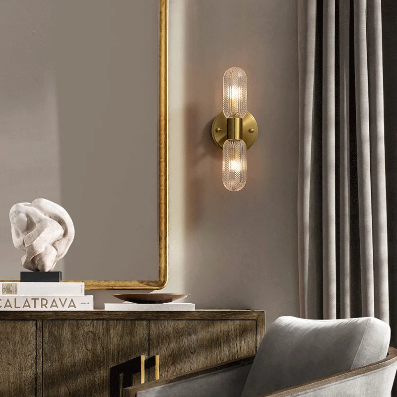 A modern living space exudes sophisticated elegance, featuring a Morsale Palermo 12" Brass Wall Light Sconce with two bulbs and textured glass covers. Adjacent is a wooden cabinet topped with books and an abstract white sculpture. A large, gray-framed mirror and gray curtains complete the refined decor.