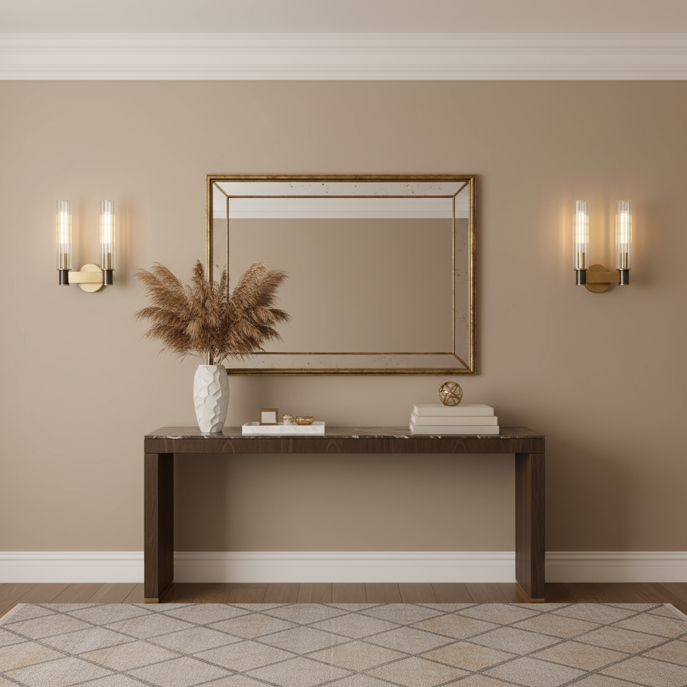 A modern entryway showcases a dark wood console with a white vase, books, and decor. On the beige wall above, a large mirror is flanked by two Morsale Palermo 2-Light Wall Sconces, with a patterned rug tying the space together.
