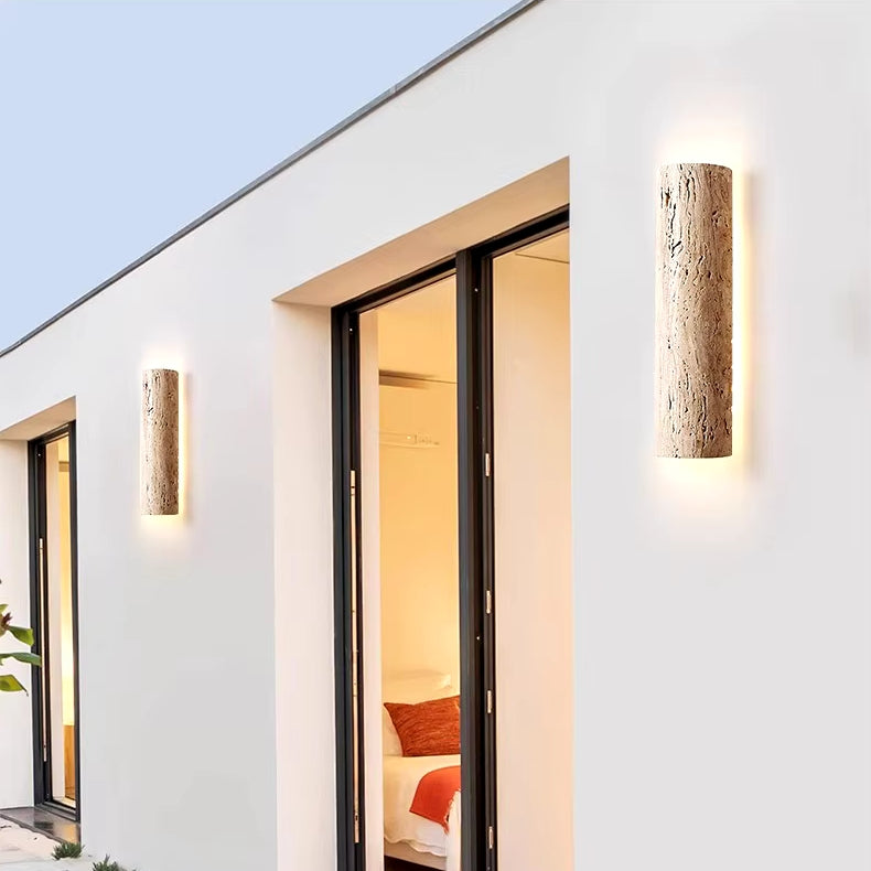 The modern building facade showcases two vertical YEBMLP IP65 Waterproof Natural Travertine Wall Lights, which are energy-efficient and emit a cozy warm glow to light up the area. Glass sliding doors open into a bedroom with white walls and a visible bed, all beneath a clear sky.