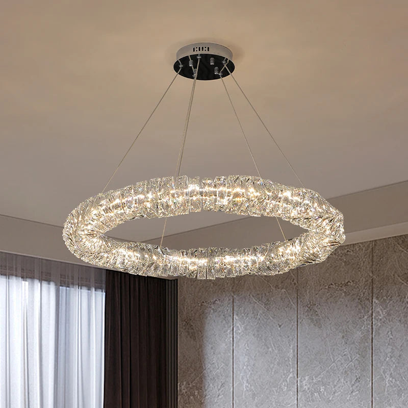 The Bacci 39" Crystal Chandelier from Morsale.com, a modern circular LED pendant with a textured crystal-like design, hangs by four wires in a stylish room featuring large windows and neutral walls.