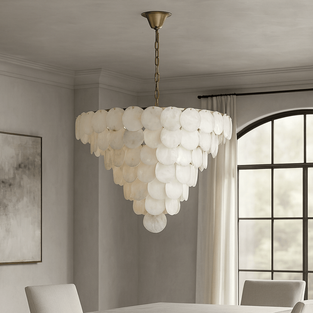 A modern dining room showcases the Porto Marble Modern Chandelier by Morsale.com, featuring overlapping round white capiz shells on a brass chain above the table. The space includes light gray walls, white curtains, and a framed abstract painting.