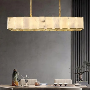 A modern dining room with a rectangular Natural Marble Ceiling Chandelier from Morsale.com hanging above a wooden table set with a green and brown vase, a small white model tree, a brown cup, and three small bowls. The room features a minimalist design with a grey wall and vertical slats, complemented by an elegant brass light fixture.