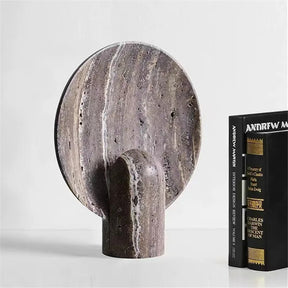A modern stone sculpture with a circular disc attached to a cylindrical base stands on a white surface. Beside it, two black books with white and gold lettering are positioned upright. The background is plain and white, creating the perfect setting for showcasing a Morsale Natural Travertine Table Lamp with adjustable brightness.